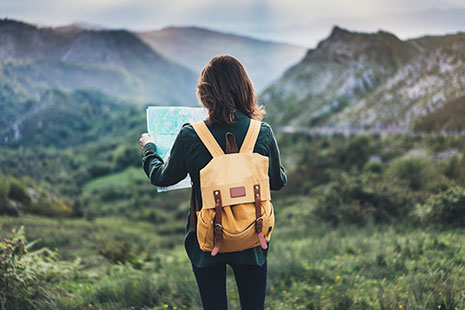 Mujer de espaldas con mochila amarilla y consultando mapa frente a montañas