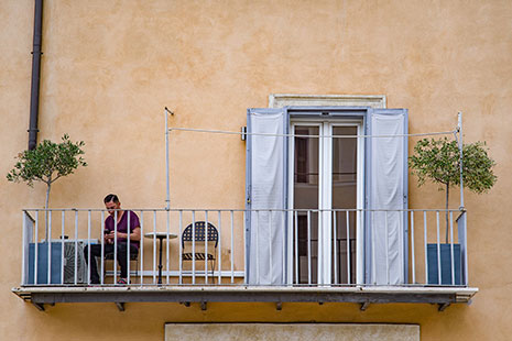 Hombre sentado en un balcón con árboles y puerta abierta