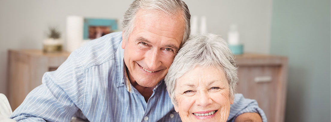 : Una pareja de ancianos posa sonriente. La mujer se encuentra sentada en un sofá gris y va vestida con una camisa blanca. El hombre detrás de ella se apoya en sus hombros con una camisa azul. De fondo una cómoda donde descansan fotos. 