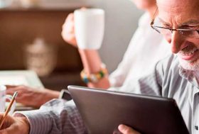 Una pareja madura sonriente se encuentra sentada en una mesa. El hombre sostiene una tablet y escribe sobre una libreta. La mujer se encuentra al fondo algo difuminada escribe en un ordenador y sostiene una taza de café