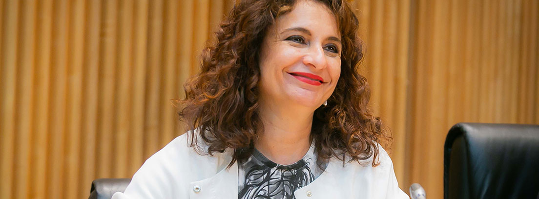 María Jesús Montero con chaqueta blanca y sonrisa sentada ante micrófono