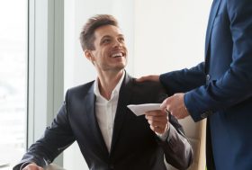 Un hombre de pie y con traje da un sobre a un trabajador feliz sentado en una mesa