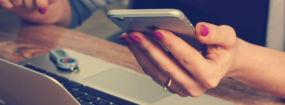 Manos de mujer sobre un ordenador portátil sujetando un móvil para pedir la vida laboral por SMS