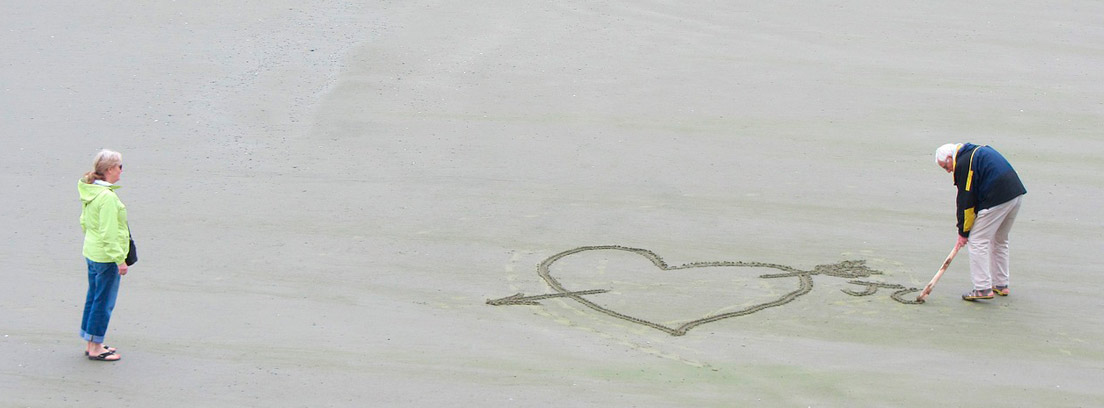 Mujer mira a hombre de pelo blanco que dibuja corazón en arena de la playa