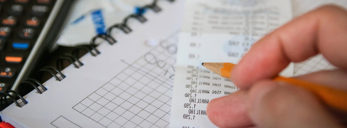 Primer plano de una mano con un lápiz sobre cuadernos, calculadora y un ticket de compra