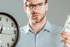 Hombre con gafas y gesto serio sujetando un reloj en una mano y unos billetes en la otra