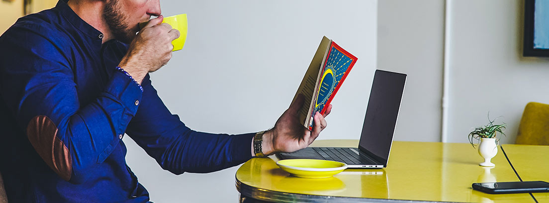 Hombre bebiendo de taza amarilla, sentado delante de ordenador portátil y mirando un libro
