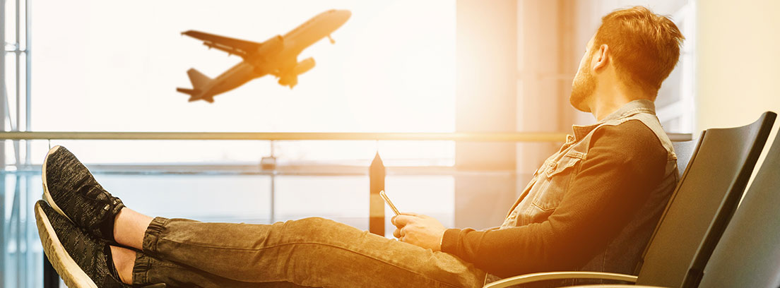 Hombre sentado con pies sobre maleta mira avión despegando