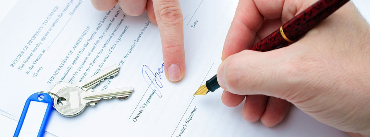Vista de las manos de dos personas firmando un contrato de alquiler y unas llaves en la mesa.