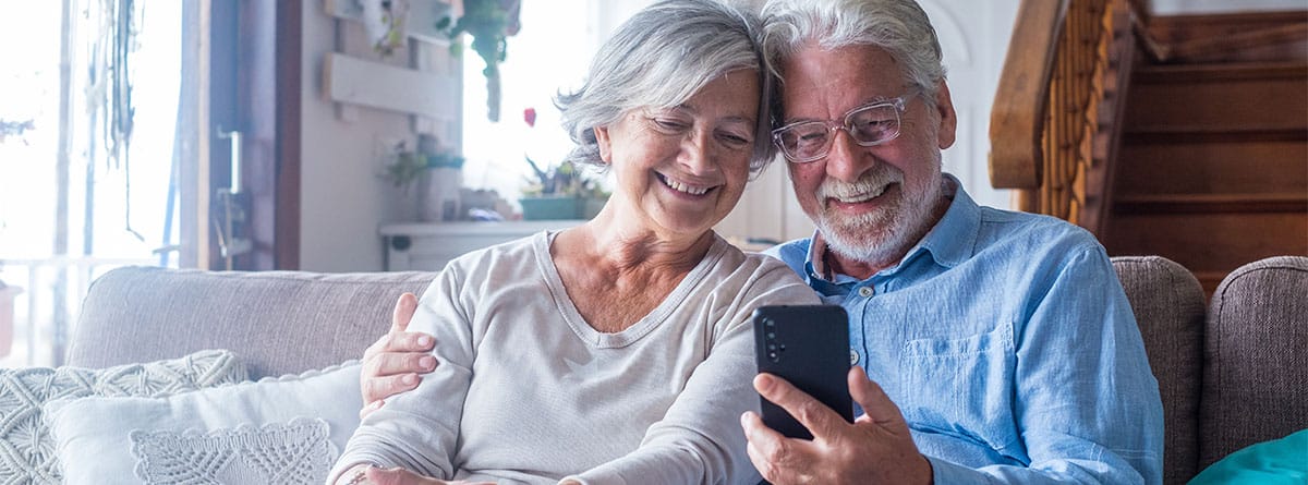 Una pareja de personas mayores sentadas en un sofá miran un móvil y sonríen.