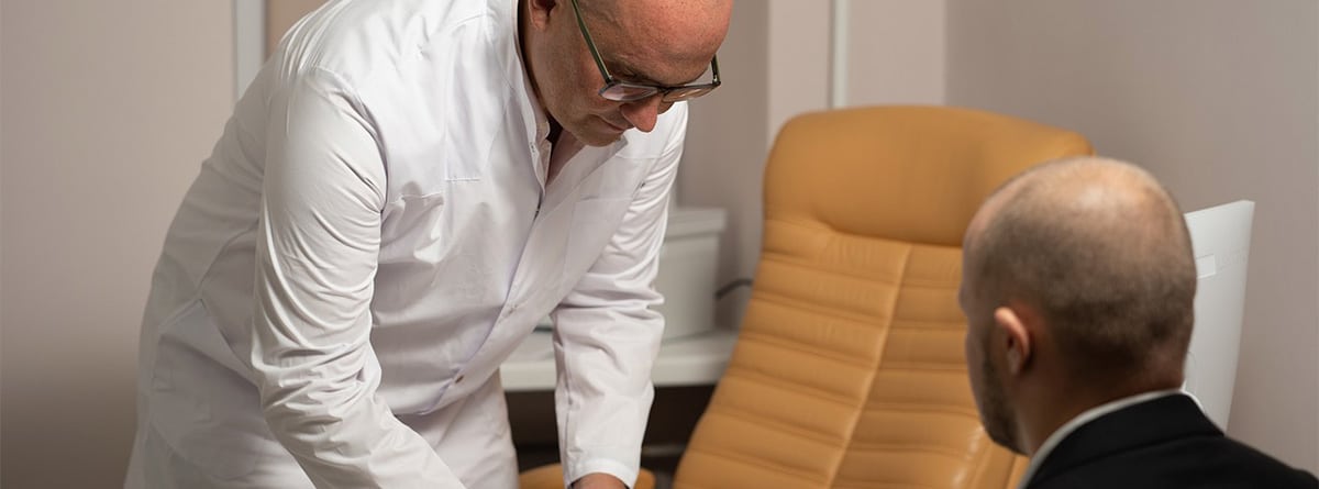 Un médico con bata blanca de pie firma un documento en presencia de otro hombre sentado.